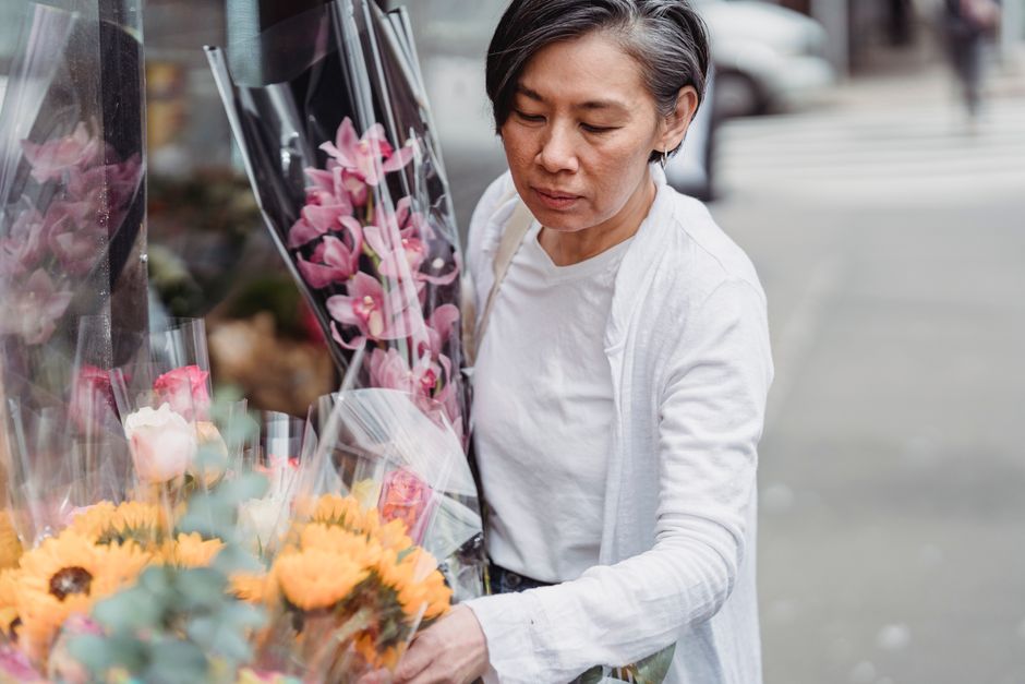 A middle-aged woman with short dark hair, wearing a white long-sleeve top and light-colored cardigan, selecting a bouquet of fresh pink orchids wrapped in clear plastic from a display of colorful flow