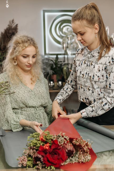 A professional florist from flowerskingsbury.co.uk is creating a floral gift arrangement, wrapping a bouquet of vibrant red roses, pink waxflowers, and green foliage with red tissue paper. The florist