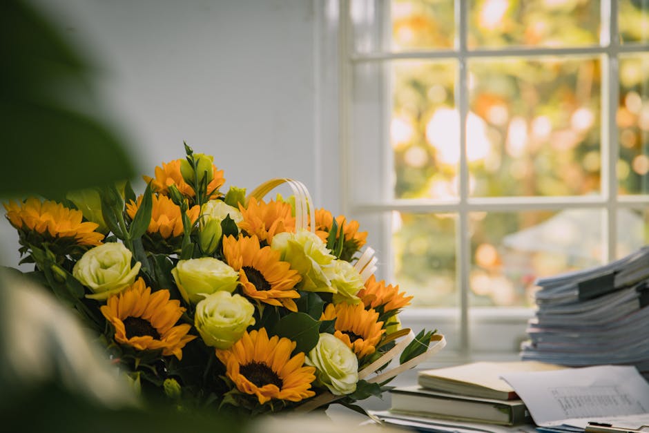 A vibrant floral bouquet featuring bright yellow sunflowers, creamy white lisianthus, and green foliage arranged in a natural, loose style with a white ribbon. The bouquet is placed on a desk near a l