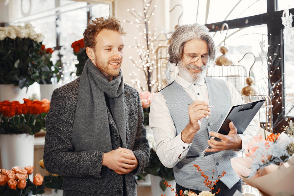 A male customer and a male florist are inside a well-lit flower shop with large windows. The florist, an older man with gray hair and a beard, wearing glasses, a white shirt, and a light gray vest, is