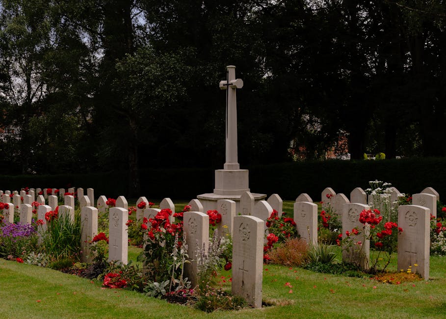 A peaceful cemetery scene featuring rows of white gravestones arranged in a semi-circle around a tall, white memorial cross. In the foreground, there are vibrant red carnations and purple flowers, wit