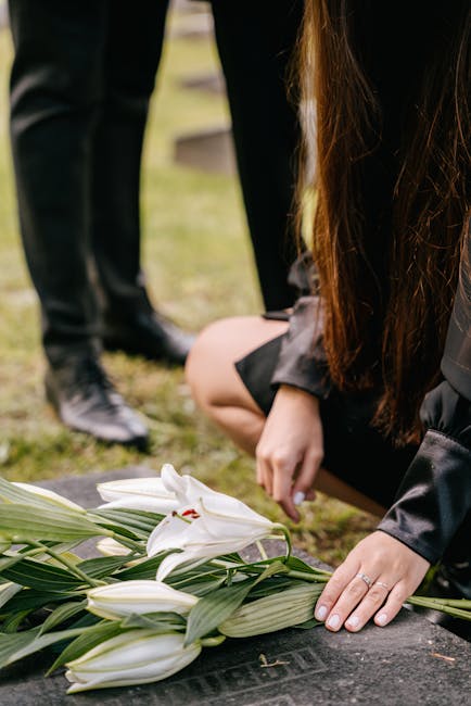 A person with long brown hair, dressed in a black jacket, is kneeling on a grassy surface placing a white lily floral arrangement on a gravestone. The bouquet features large white lilies with open pet