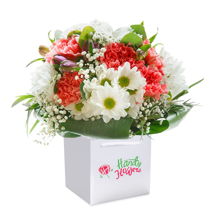 Set against a luminous, pure white backdrop, this premium bouquet stands tall in a refined square gift bag, its elegance unmistakably suited for Kingsbury's refined taste. The arrangement showcases a harmonious blend of coral-pink and peach carnations-each petal richly textured and gently ruffled, their soft hues interlaced with vivacious red tones. Scattered among these are crisp white daisy chrysanthemums, each flower boasting a vivid chartreuse-yellow center that sparkles with vitality. Wisps of white baby's breath meander throughout, imparting a barely-there lightness that invokes the freshness of spring mornings in Eton Grove Park. Deep green foliage provides a bold, glossy frame, encircling the bouquet with understated grace and ensuring every bloom is displayed to enchanting effect. Along the left edge, several unopened buds tinged with subtle purples and pinks offer both visual mystery and a promise of more beauty to open in days ahead. The bouquet sits elegantly within a white bag, the handles sturdy and well-finished for easy gifting or seamless local delivery. The branding mark is present but discreet, reinforcing the feeling of an exclusive, personal gesture. Illuminated by soft, even lighting, every nuance of colour and texture is exalted, making the floral artistry of this arrangement unmistakable. Ideal for gifting-whether for a birthday, milestone, or as a spontaneous act of affection-this bouquet is a true celebration of Kingsbury's attention to quality, freshness, and local connection.