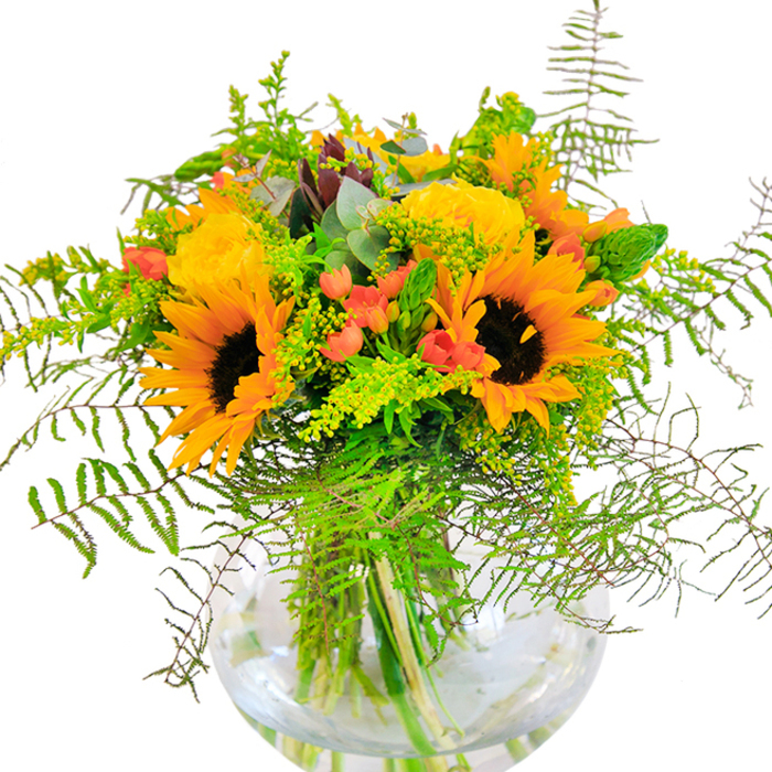 A sun-drenched celebration of colour and natural shape, this opulent bouquet features the boldest summer sunflowers, their radiant yellow petals cupping dark, earthy centers that mimic the rich gardens of Kingsbury in midsummer. Interlaced among these giants, yellow garden roses offer a classic twist; their tightly wound petals unfold delicately, recalling velvet cushions and golden sunlight filtering through Forrest Road in the afternoon. The arrangement is playfully interrupted by sprigs of glossy red Hypericum berries, little bursts of scarlet glinting amidst a sea of greens and golds-each one catching the light much like berries along the wooded paths of Fryent Way. Cascading ferns and wispy sprigs of filler foliage lend airy movement, their soft texture inviting touch and their delicate hues harmonising with the composition's gentle rusticity. Light green goldenrod nods between larger flowers, its minuscule yellow blossoms adding whispers of texture and late summer fullness. Bluish eucalyptus leaves introduce a tender, cool note, tucked among the brightness and balancing the palette. The entire bouquet is cradled in a sparkling, round glass vase, where the waterline magnifies the intricate crisscross of fresh stems. The result is a luminous, locally inspired centrepiece ideal for birthdays, new beginnings, or simply adding a sense of joy and community spirit to a Kingsbury living space.